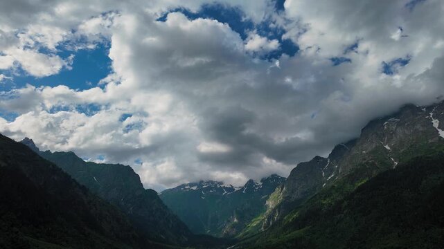 Majestic view of white clouds moving over a green mountain valley with snow-capped peaks against a bright blue sky, creating a stunning and dynamic natural landscape Georgia Svaneti