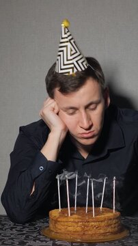 Bored and sleepy man wearing a party hat celebrating a solitary birthday, listlessly blowing out the candles on his cake before looking at the camera with a sad and lonely expression