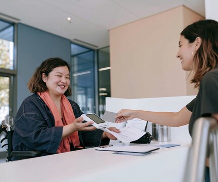 East Asian woman in a wheelchair staffing a fundraising stall, smiling while processing a donation with a blank card reader for corporate giving and accessible workplace campaigns
