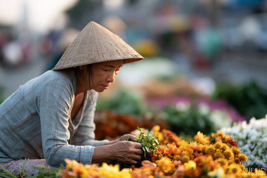 Woman in conical hat tends to colorful flowers. Her hands gently sort through blooms in market light. Soft focus background suggests bustling street scene. She focuses intently
