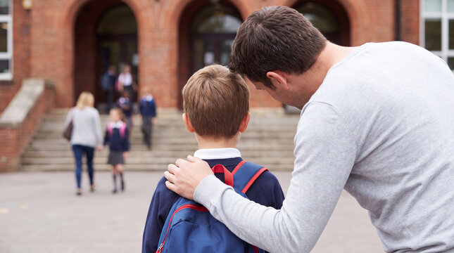 Father comforts young boy with backpack outside school building as children walk up steps in the background during school day