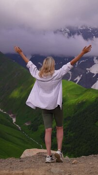 Blonde woman standing on the edge of a cliff, raising her arms in a sign of freedom and enjoying a breathtaking view of a green valley and snow-capped mountains, partially covered in clouds in Georgia