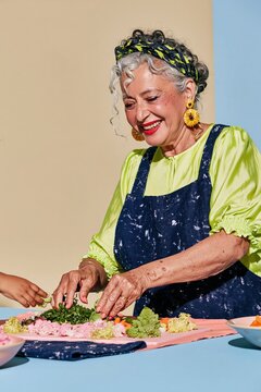 South Asian grandmother arranging fresh herbs and crudites on a pale board, featuring weathered hands and copy space for hosting and care