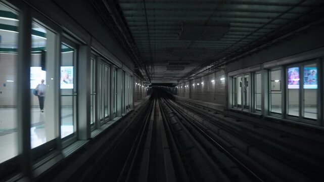 Front train perspective rides past glass station platform screen doors before accelerating deep into a dark curved subterranean subway tunnel