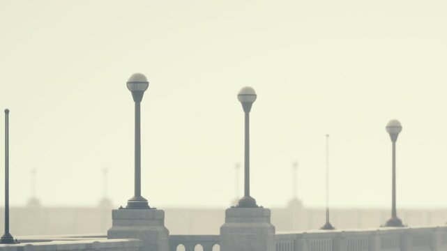 Light posts stand on a walkway near the water. The scene is set in the early morning with a soft light. Few people are present, creating a quiet atmosphere by the water.