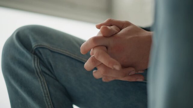 Close up of hands clasped together resting on lap with blue jeans visible soft natural light indoor setting closeup lighting texture detail factual still