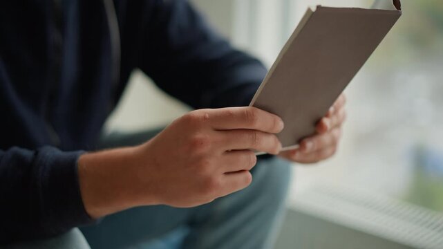 Close up of person holding and examining open notebook with both hands focusing on interaction between hand positions and book orientation. scene is set indoors near window in soft natural