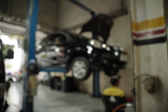 Blurred view of an auto repair shop featuring a car being lifted on a hoist. The car's hood is open, and various tools are visible nearby. The image has been blurred for use as a background.