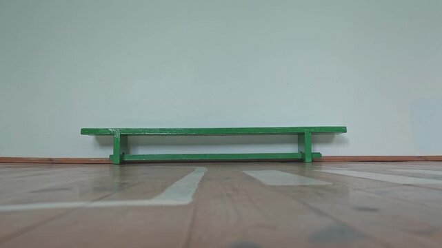 Single miniature green wooden bench standing alone in an empty room. Minimalist scene with a wooden floor and a plain white wall background