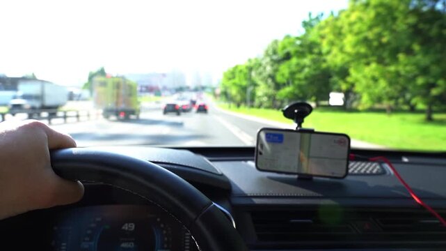 Hand grips steering wheel during drive. Phone mounted on dashboard shows map. Road ahead with cars and green trees. Driver focuses on route, avoiding traffic. Ideal for travel, navigation