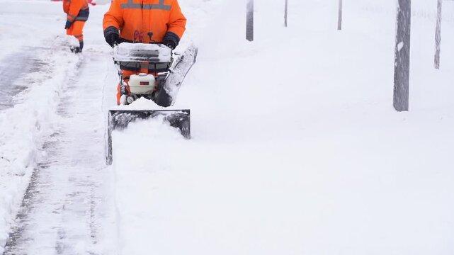 Worker clears snow with mechanical snowblower. Snow covers streets and utility poles. Worker wears bright orange uniform. Snowblower pushes snow sideways. Worker moves steadily forward