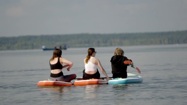 Three women paddle serene lake in kayaks. One paddles vigorously, others glide calmly. Trees line distant shore under soft sky. Sunlight glints gently on calm water surface