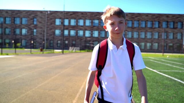 Young boy walks toward camera with backpack. School building looms behind him on sunny day. He holds books, looking calm and focused. Ideal for scenes about school life or daily routines
