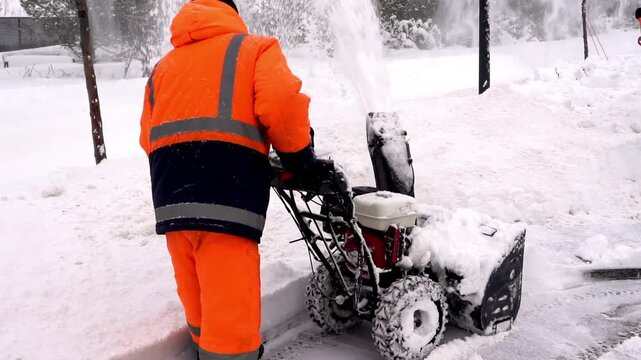 Worker clears snow with snowblower in bright orange suit. Snow flies as machine moves through deep winter accumulation. Worker focuses on task, shoulders showing effort in cold environment