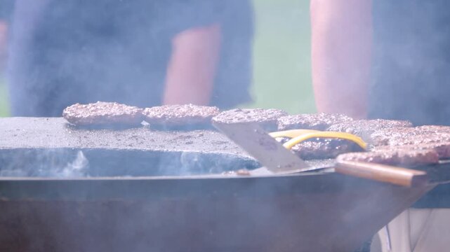 Smoke curls from sizzling patties on a grill. Hands move a spatula, turning meat with care. Grass and sky blur behind the cooking action. Outdoor gatherings feel warm and casual