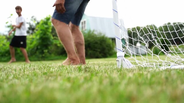 Child kicks ball toward goal on grassy field. Another child runs after the ball. Goalpost stands in soft green grass. Scene feels playful and carefree. Ideal for sports, family, or outdoor fun