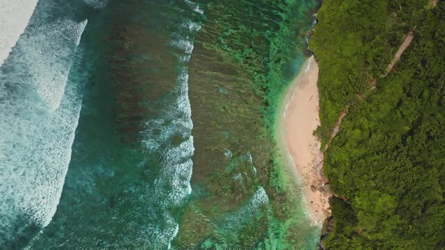 Bukit Peninsula coastline featuring turquoise ocean waves crashing on a pristine sandy beach next to a vibrant green tropical cliff, creating a serene and idyllic island escape. Aerial top down view