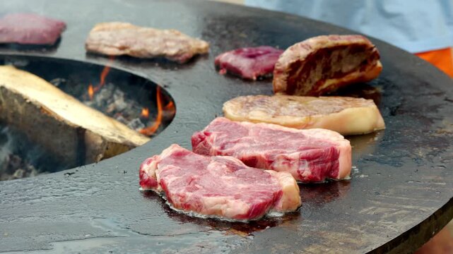 Wood fires crackle beneath sizzling steaks. Meat sizzles on hot, flat iron grills. Flames lick edges of cooking cuts. Cooking smoke drifts upward gently. Crisp, juicy meat cooks under heat