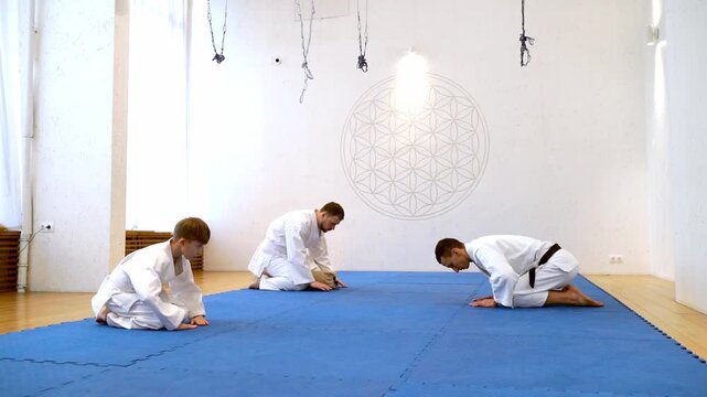 Three martial artists in white uniforms sit cross-legged on a blue mat. One bows low, hands pressed to ground, showing respect. The room holds calm, focused energy, with minimal decor