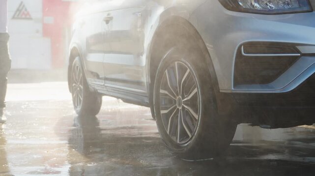 Water sprays over gleaming vehicle's side. Sunlight glints off wet chrome wheels. Person stands nearby, focused on cleaning. Ideal for automotive, service, or maintenance themes