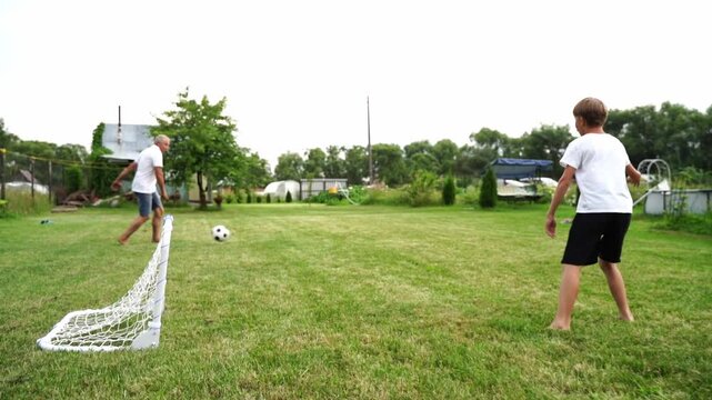 Sunny day. Two people play soccer barefoot on grass. White goal stands ready. One runs to kick ball. Other watches from distance. Casual, joyful movement