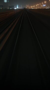 Front train perspective advances along elevated railway track toward a rising section, passing highway lights and distant buildings at night
