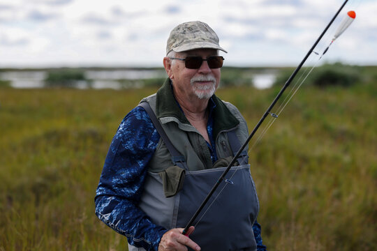 Mature man wearing waders and camouflage cap holds fishing rod with float, smiling in Alaska&rsquo;s open wilderness under cloudy sky, enjoying peaceful outdoor recreation near water.