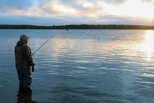 Fisherman in waders watching the sunrise in Alaska. He is holding fishing pole wearing camouflage.