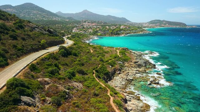 Ile-Rousse Corsica coastal road drone overflight driving cars scenic seaside drive. Aerial view of rugged coastline, turquoise sea waves, distant island islet panorama France travel 4K.