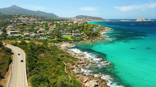 L Ile Rousse coastal road tracking shot with cars and yellow ferry in Corsica France. Aerial flyover of rocky coast pink luxury villas turquoise pools 4K
