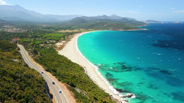 Aerial drone tracking shot along scenic coastal road and long Lozari sand beach in Corsica France. Turquoise Mediterranean sea near L'Ile Rousse 4K