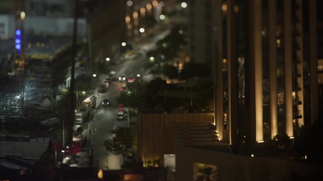 Cars move through traffic along a city street at night, passing illuminated buildings and streetlights