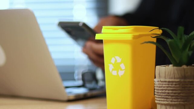 Yellow recycling bin with recycle symbol on an office desk with an unrecognizable person working on a laptop promoting environmental awareness and sustainability