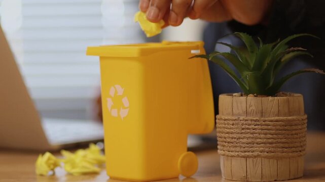 Hand throwing crumpled yellow paper into a small yellow recycling bin on an office desk, showcasing eco friendly waste management and environmental responsibility
