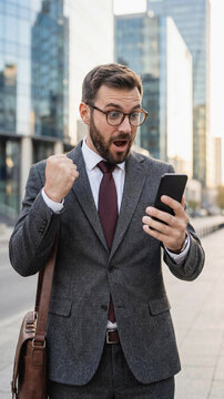 Excited young businessman in a grey suit and glasses reading good news on his smartphone outdoors, celebrating success or victory with joy in a modern city environment