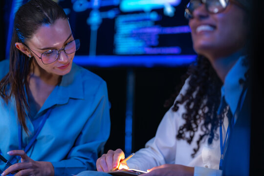 An Asian developer and a bespectacled coder two teammates talk closely while seated in a modern technology office bathed in blue light, reflecting diversity and innovation in a startup