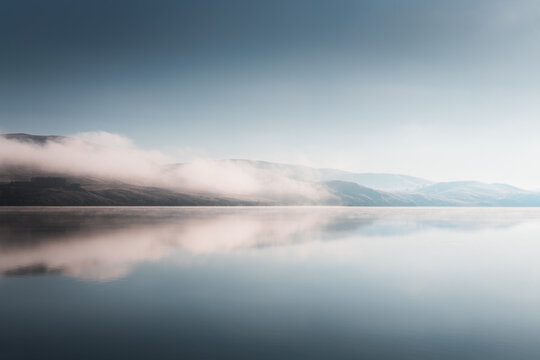 Beautiful lake at foggy sunrise. Mountains with clouds are reflected in the calm water. Autumn landscape