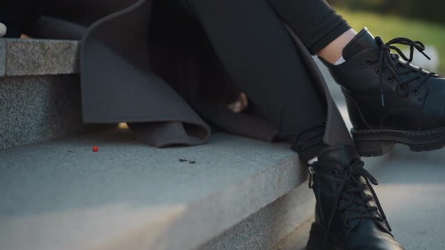 Close up of woman legs and feet in black combat boots resting on stone steps with soft lighting. dark coat drapes over lap while nearby sits white mesh bag containing oranges and red berries