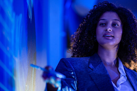 Businesswoman with curly hair outlines the roadmap with open hands rim lit in gold while speaking beside a deep blue wall in a modern briefing theatre glowing with interactive data graphics