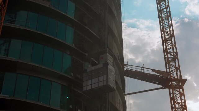 Construction elevator travels along an unfinished high rise beside a tower crane, with curved concrete floors and glass panels under a cloudy sky