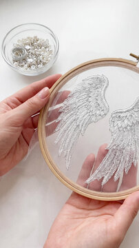 Female hands holding embroidery hoop with delicate silver wings stitched on mesh fabric. Creative process of making bridal handmade accessories with pearls and sequins in glass bowl on white table.