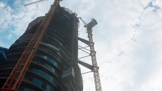 Tower cranes stand beside an unfinished high rise, as the camera tilts upward along curved concrete floors and glass panels