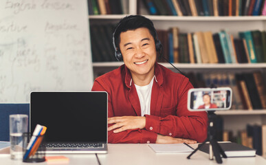 Fototapeta premium Smiling Asian Male Educator Engaging With Students Online From Home Office Using Laptop, Wearing Headset. Focused On Teaching In A Comfortable Environment, Promoting Online Learning Concept.