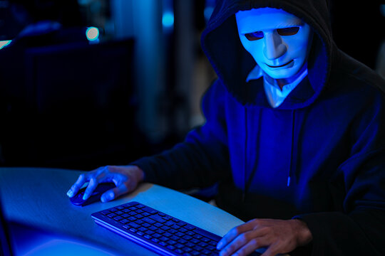 An anonymous cyber criminal wearing a white mask and black hoodie typing on a glowing keyboard to launch a malicious network database ransomware attack.