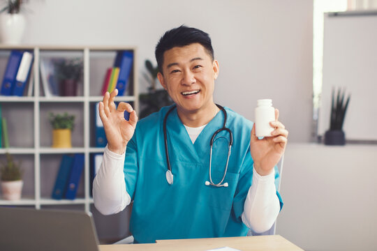 Smiling Asian Male Doctor In His 30s Showing A Pill Bottle And Making Okay Gesture In His Office While Sitting At Desk. Promoting Medicine And Health Care In A Professional Environment.