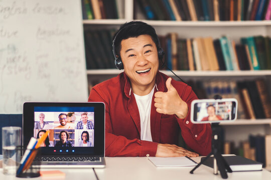 Smiling Asian Male Instructor Giving Thumbs Up While Teaching Online Class With Students On Laptop, Sitting At Desk In Modern Office Setting, Engaging Emotions, Business Concept.