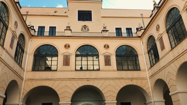 Tilt up shot revealing the grand classical facade and arched loggia of an elegant Italian palazzo inner courtyard featuring historic frescoes, ornate windows, and a bright blue sky.

