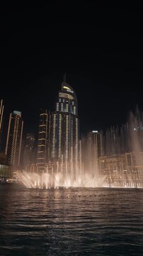 Dubai Fountain water jets rise in slow motion before Address Downtown at night, reflecting across Burj Lake