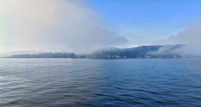 Horizontal view of misty island coastline shrouded in thick fog over calm blue ocean water