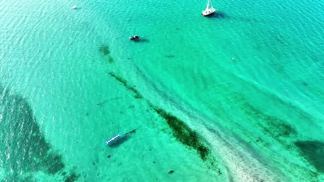 Aerial drone video of turquoise coastal water, shallow sandbanks, dark seaweed patterns and boats gliding across a crystal-clear shoreline